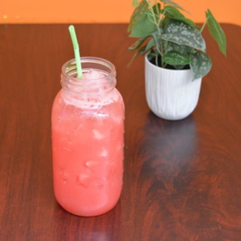 Pink drink in a mason jar with a green straw, next to a potted plant on a wooden table.