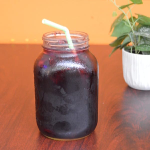 A frosty mason jar of dark red agua fresca with a green straw on a wooden table.